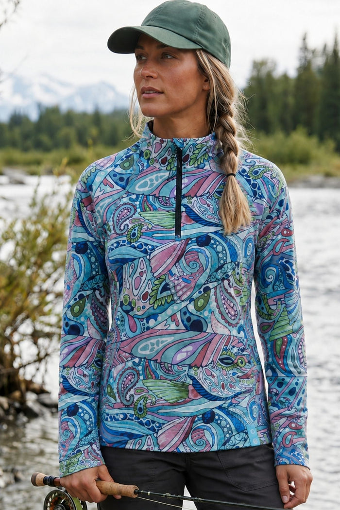 Woman wearing a colorful patterned long-sleeve shirt by a body of water with mountains in the background.