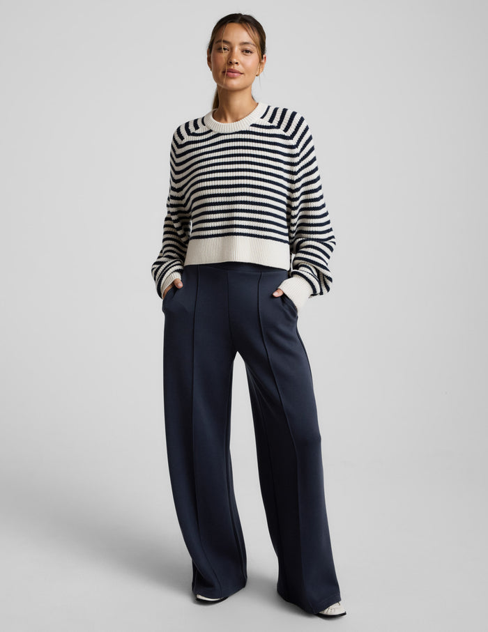 Woman wearing a white and blue striped hi lo sweater and blue pants against a light gray background.