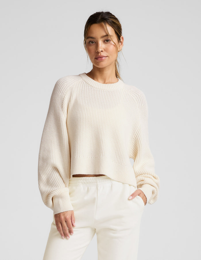 Woman wearing a beige hi lo sweater against a light gray background.