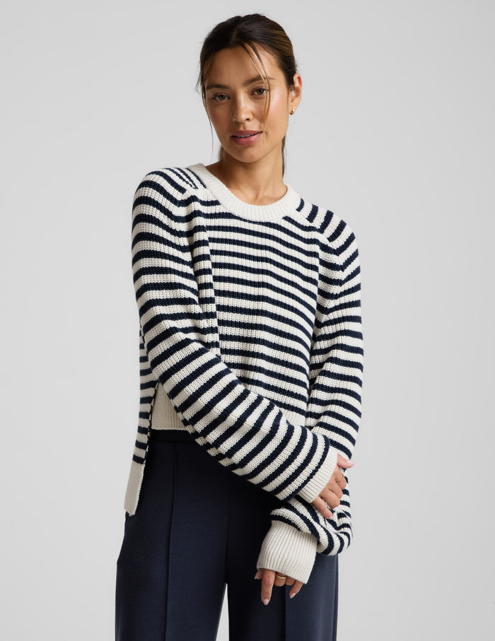 Woman wearing a white and blue striped hi lo sweater and blue pants against a light gray background.