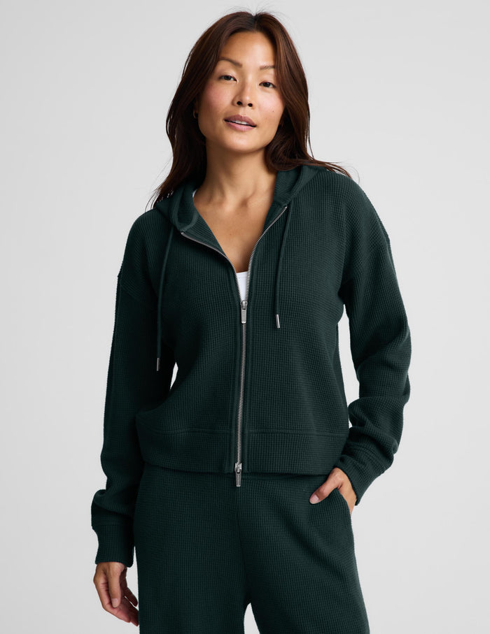 A woman modeling a dark forest green ribbed knit zip-up hoodie and matching sweatpants against a light gray studio background.