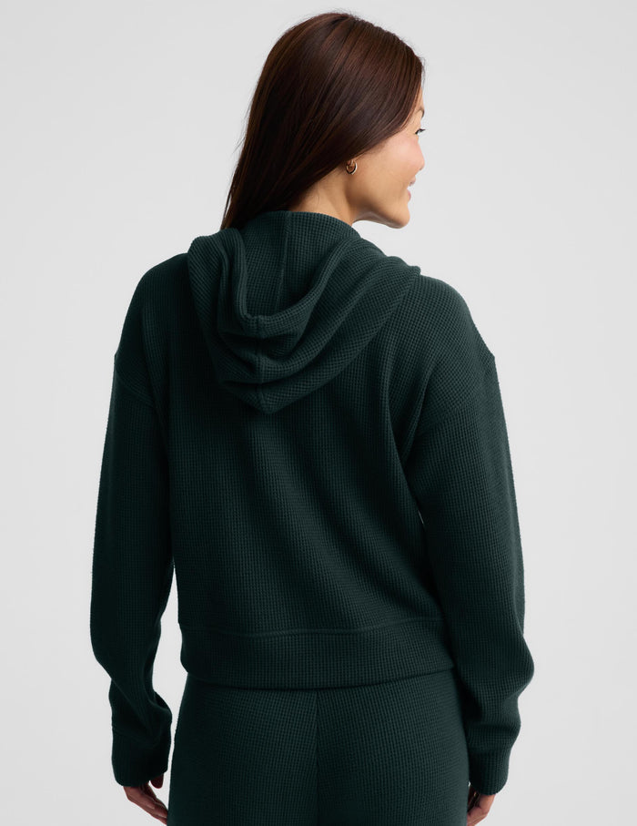 A woman modeling a dark forest green ribbed knit zip-up hoodie and matching sweatpants against a light gray studio background.