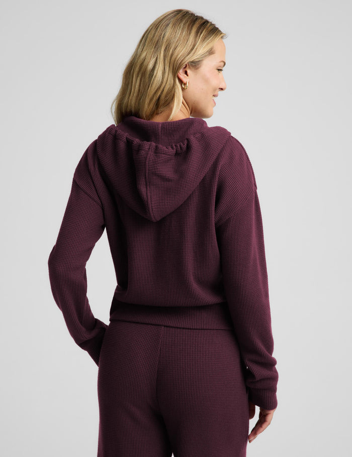A woman modeling a matching burgundy waffle-knit zip-up hoodie and sweatpants set against a light gray studio background.