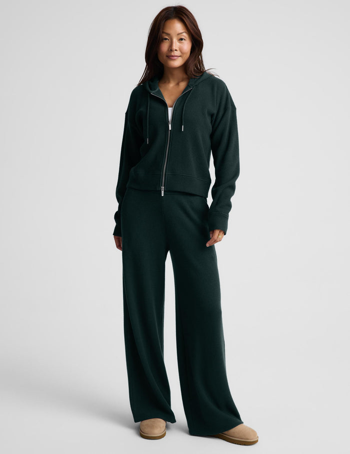A woman modeling a dark forest green ribbed knit zip-up hoodie and matching sweatpants against a light gray studio background.