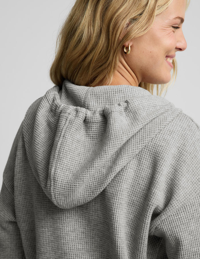 A close-up shot of a woman wearing a gray waffle-knit hooded sweatshirt, shown from a side-back angle against a light gray studio background.