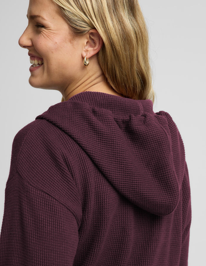 A woman modeling a matching burgundy waffle-knit zip-up hoodie and sweatpants set against a light gray studio background.
