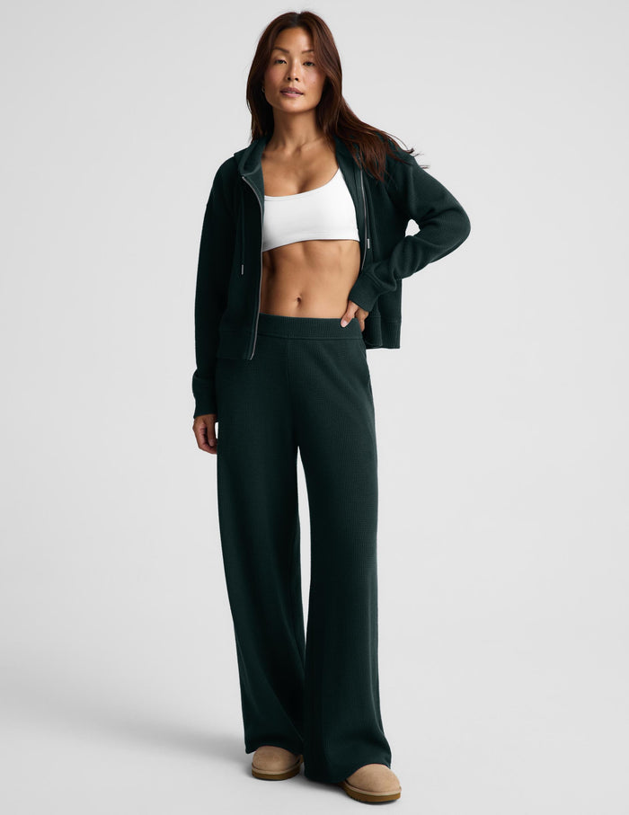 A woman modeling a dark forest green zip-up hoodie and matching wide-leg sweatpants, styled with a white sports bra and tan slip-on shoes against a light gray studio background.
