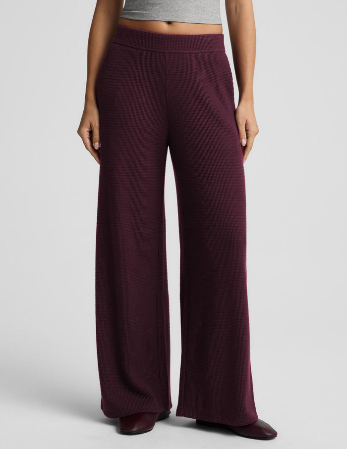 A model wearing a pair of wide-leg knit pants in a deep burgundy, paired with a gray crop top and dark brown shoes, against a light gray studio background.