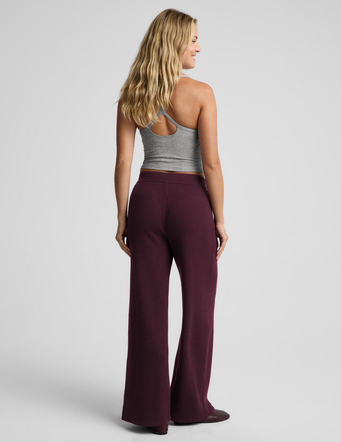 A woman shown from the back wearing high-waisted, wide-leg lounge pants in a deep burgundy/maroon color, paired with a gray racerback tank top, against a light gray studio background.