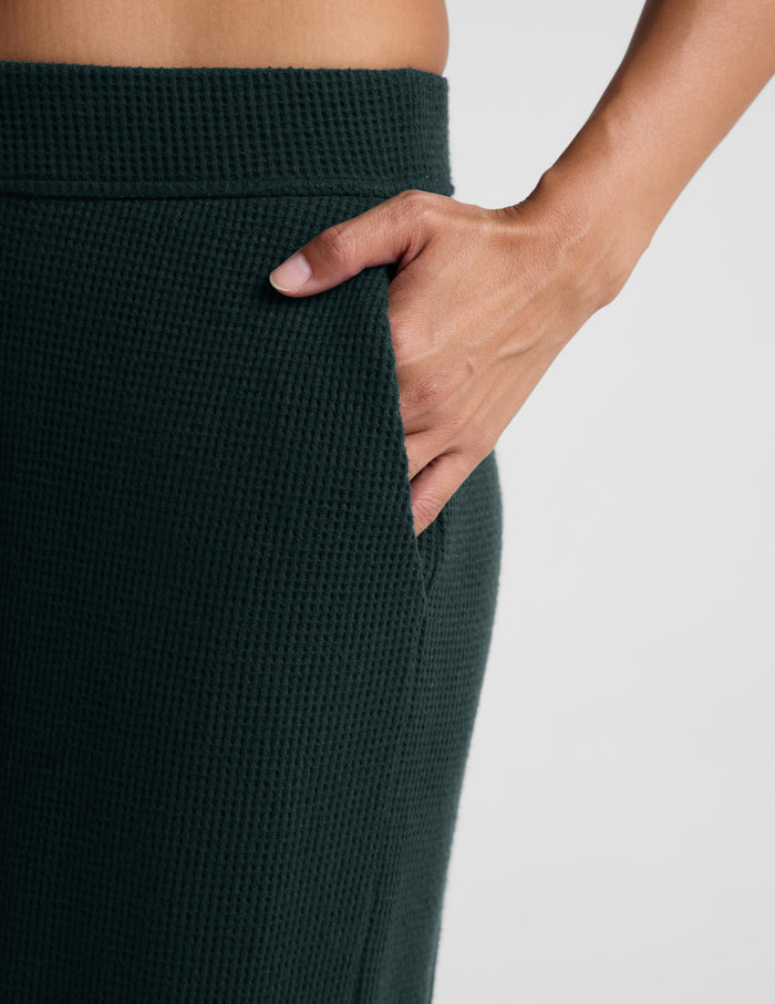 Close-up of a person wearing dark forest green waffle-knit pants, showing a hand tucked into a side pocket against a light gray background.
