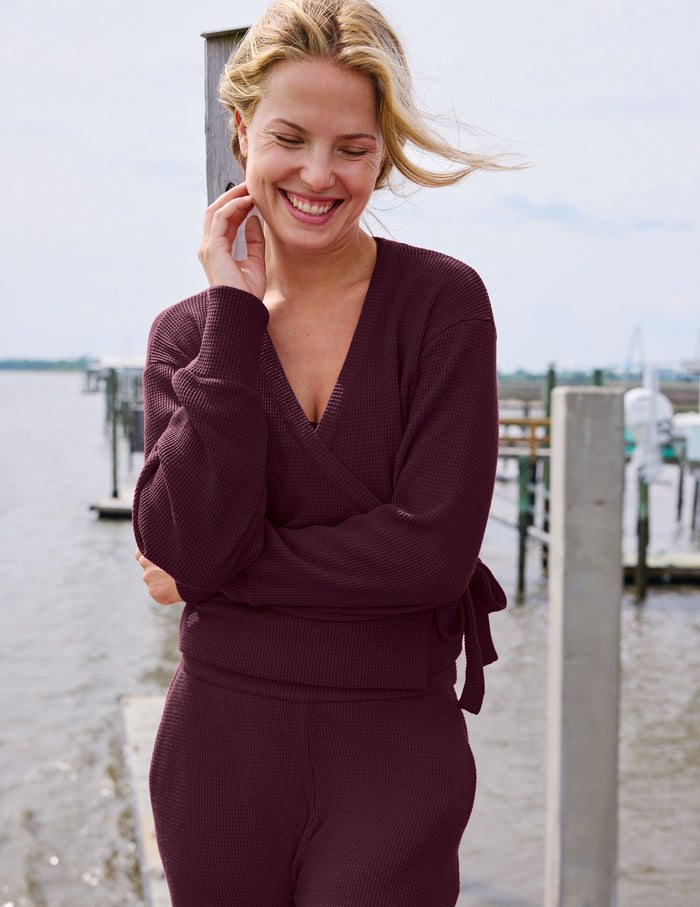A woman smiling outdoors near a waterfront dock, wearing a matching burgundy knit wrap top and pants set.