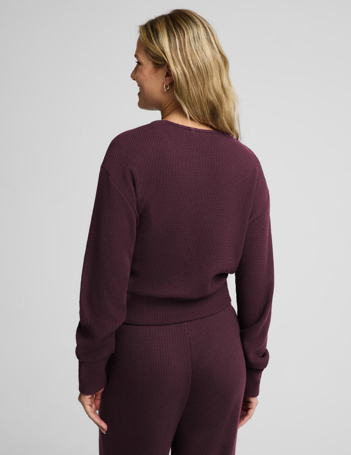 A smiling blonde woman wearing a matching deep plum purple ribbed knit loungewear set, featuring a wrap-style cardigan with a tie belt and coordinating pants, against a solid light gray background.
