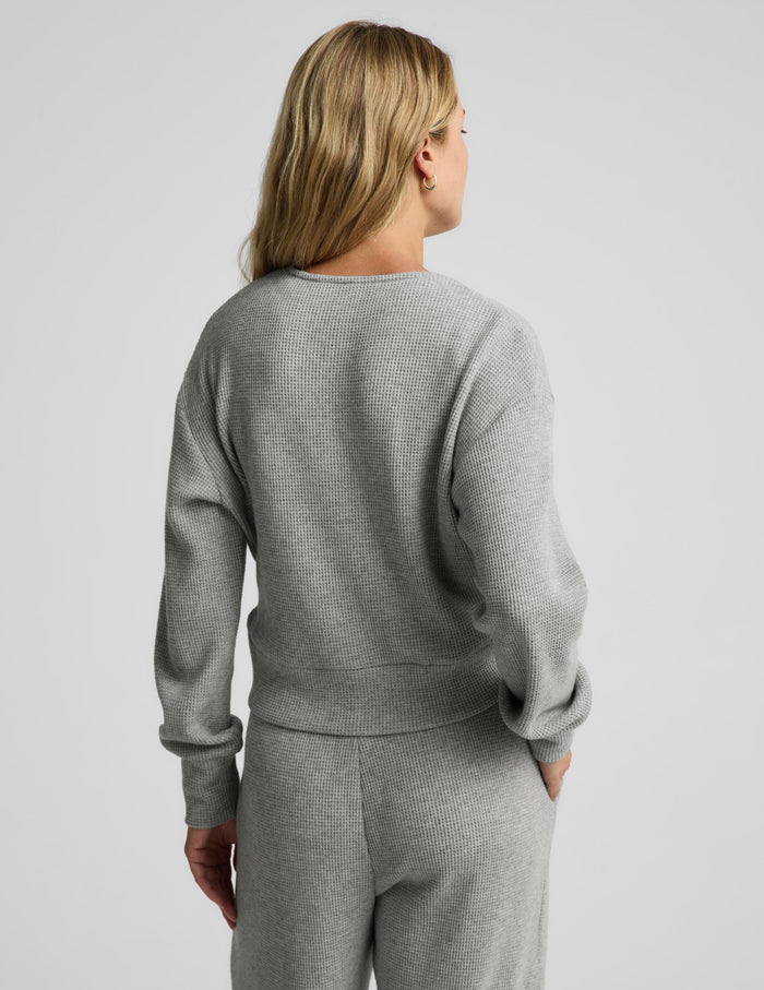 A woman modeling a light gray knit wrap cardigan and matching lounge pants against a plain light gray studio background.