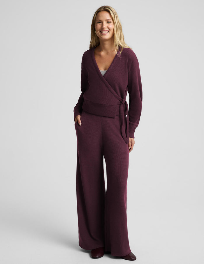A smiling blonde woman wearing a matching deep plum purple ribbed knit loungewear set, featuring a wrap-style cardigan with a tie belt and coordinating pants, against a solid light gray background.