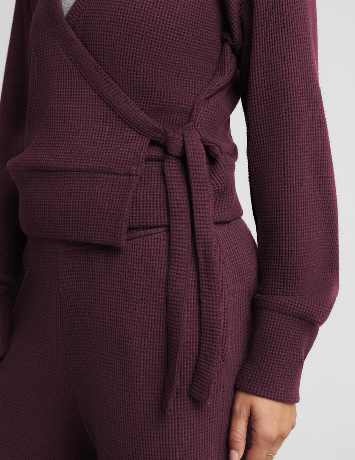 A blonde woman wearing a matching deep plum purple ribbed knit loungewear set, featuring a wrap-style cardigan with a tie belt and coordinating pants, against a solid light gray background.