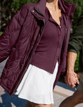 A person wearing a quilted plum-burgundy jacket over a matching button-front top and a white skirt, photographed outdoors while holding hands with another person.