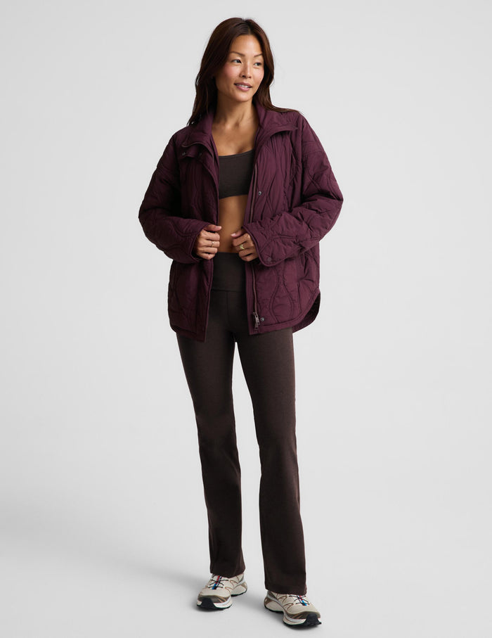 A woman modeling a quilted jacket in a deep burgundy purple color, standing against a solid light gray background.