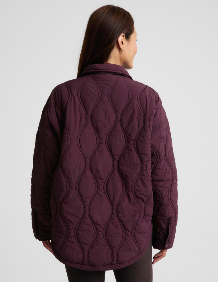 A woman modeling a quilted jacket in a deep burgundy purple color, standing against a solid light gray background.