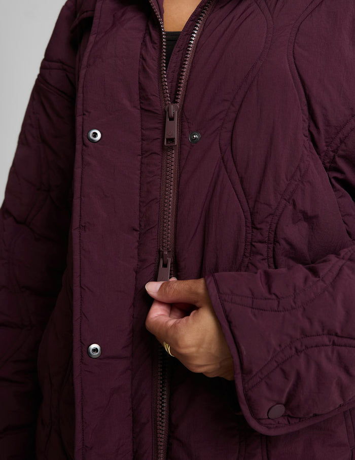A woman modeling a quilted jacket in a deep burgundy purple color, standing against a solid light gray background.