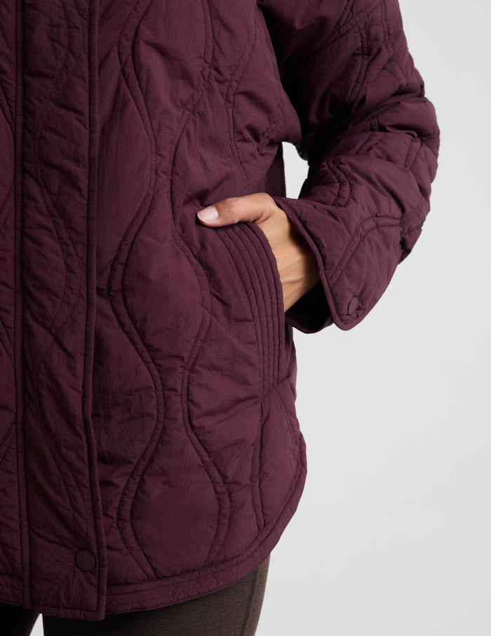 A woman modeling a quilted jacket in a deep burgundy purple color, standing against a solid light gray background.