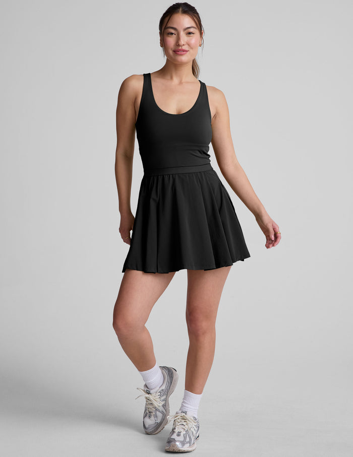 black mini dress in our stretch woven fabric with stitch down front pleats, a scoop front neckline​, open back detail​, bodice in performance stretch fabric​, and skirt side vents for easy pocket access​.