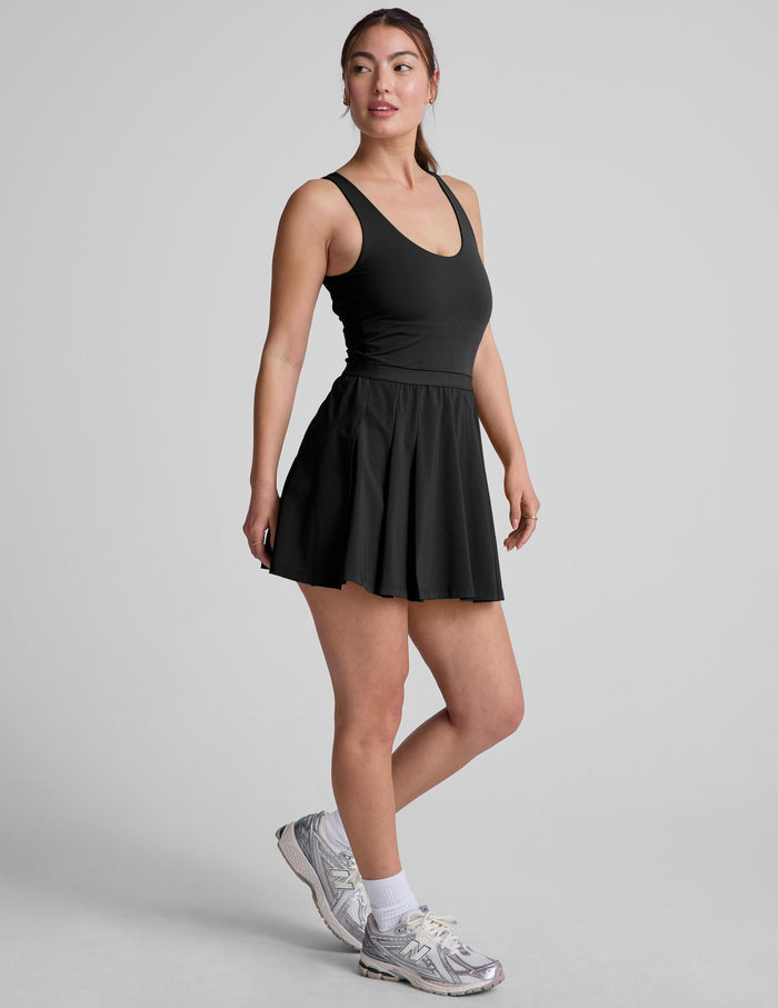 black mini dress in our stretch woven fabric with stitch down front pleats, a scoop front neckline​, open back detail​, bodice in performance stretch fabric​, and skirt side vents for easy pocket access​.