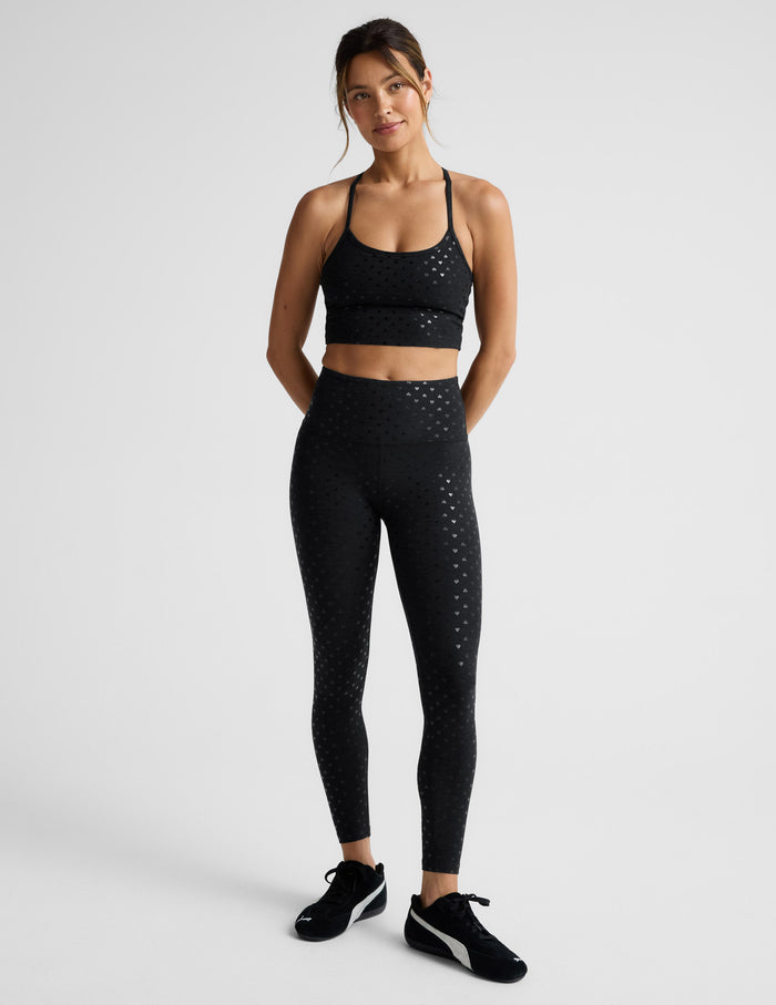 Woman wearing a black cropped tank with hearts print and leggings set on a light gray background.