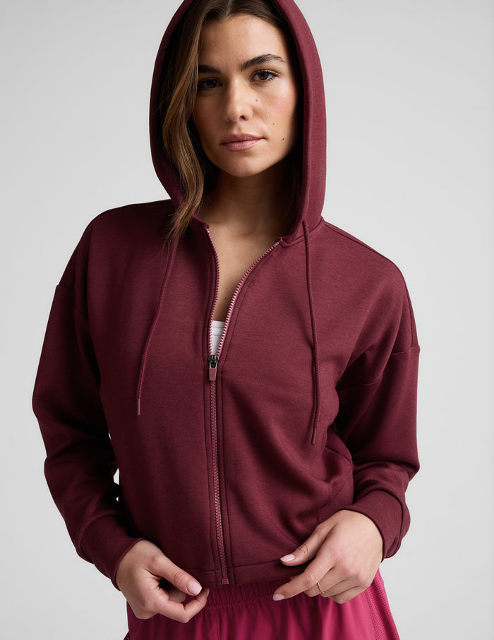 Woman wearing a burgundy zip-up sweatshirt and shorts against a plain background.