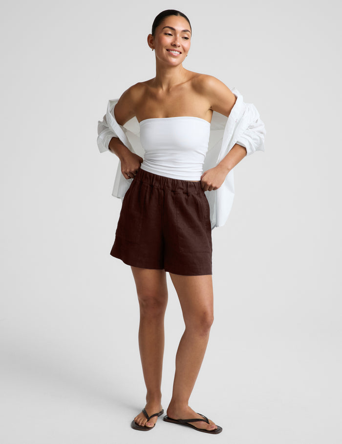 Woman wearing a white strapless tank and brown linen shorts against a plain background.