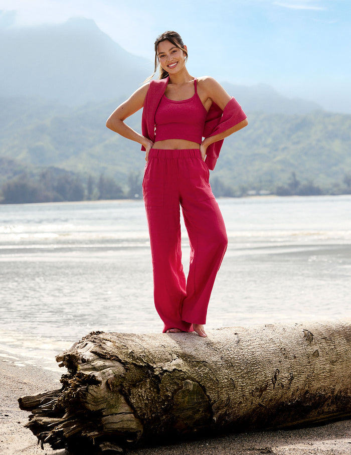 Woman wearing a red cropped tank and linen pants standing on a tree branch in front of the ocean.