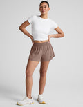 Model in white crop top wearing brown Solemate 2.5" Short with elastic waist and relaxed fit, standing against gray backdrop.