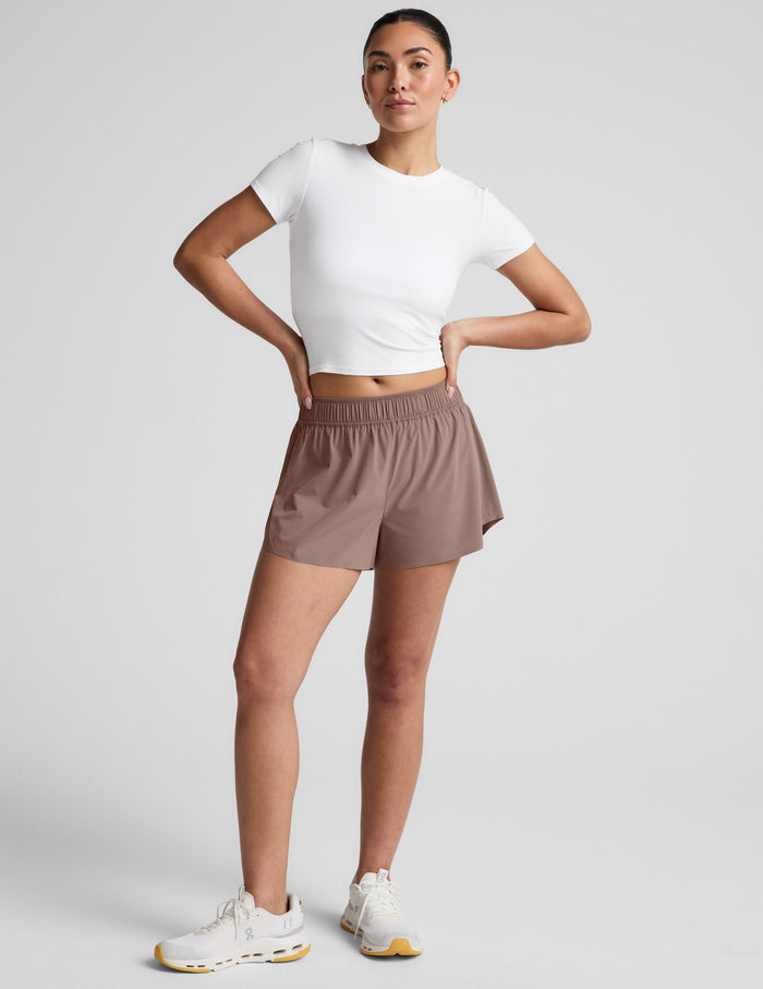 Model in white crop top wearing brown Solemate 2.5" Short with elastic waist and relaxed fit, standing against gray backdrop.