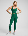 Woman wearing a green cropped v-neck tank top and green midi length leggings with a front crossover on waistband against a plain studio background.