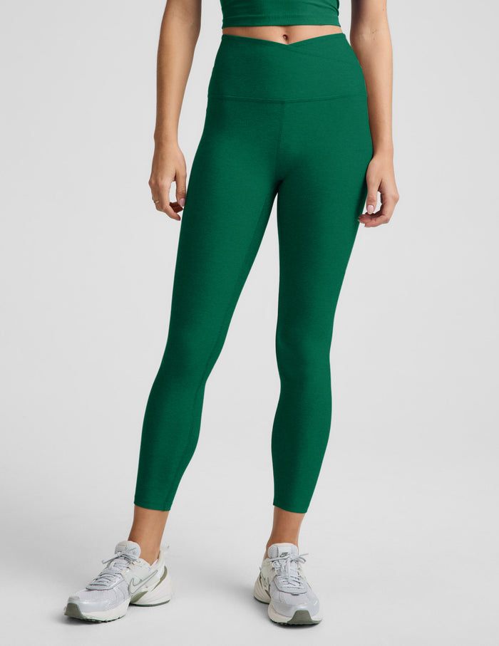 Woman wearing a green cropped v-neck tank top and green midi length leggings with a front crossover on waistband against a plain studio background.