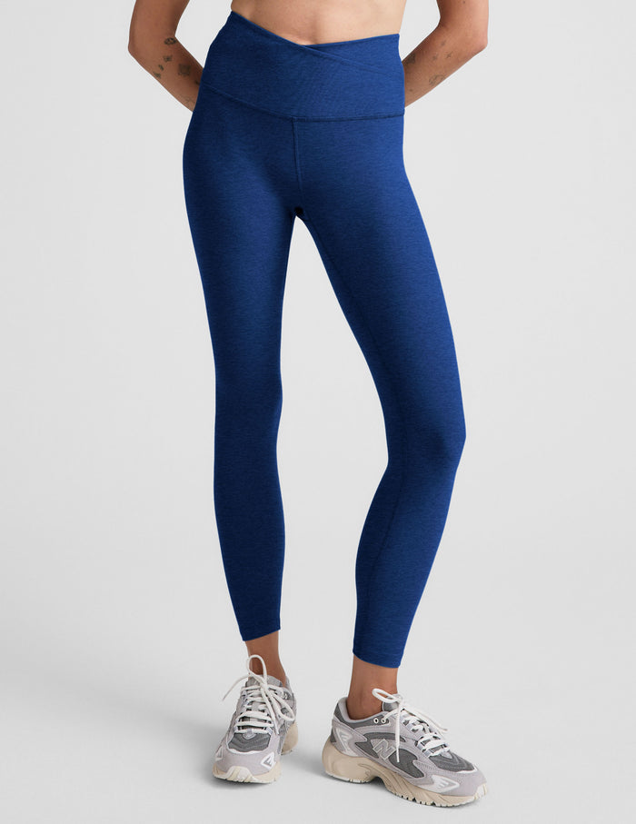 Woman wearing blue high-waisted midi leggings with a crossover front waistband against a plain background.