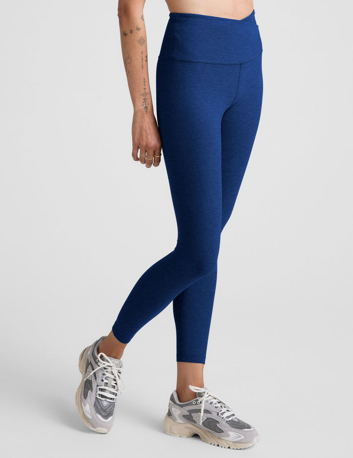Woman wearing blue high-waisted midi leggings with a crossover front waistband against a plain background.
