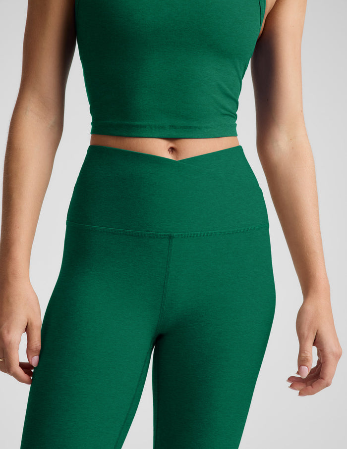 Woman wearing a green cropped v-neck tank top and green midi length leggings with a front crossover on waistband against a plain studio background.