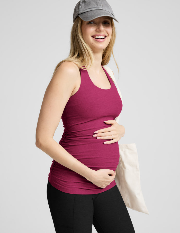 Pregnant woman wearing a pink tank top and black leggings, holding her belly against a plain background.