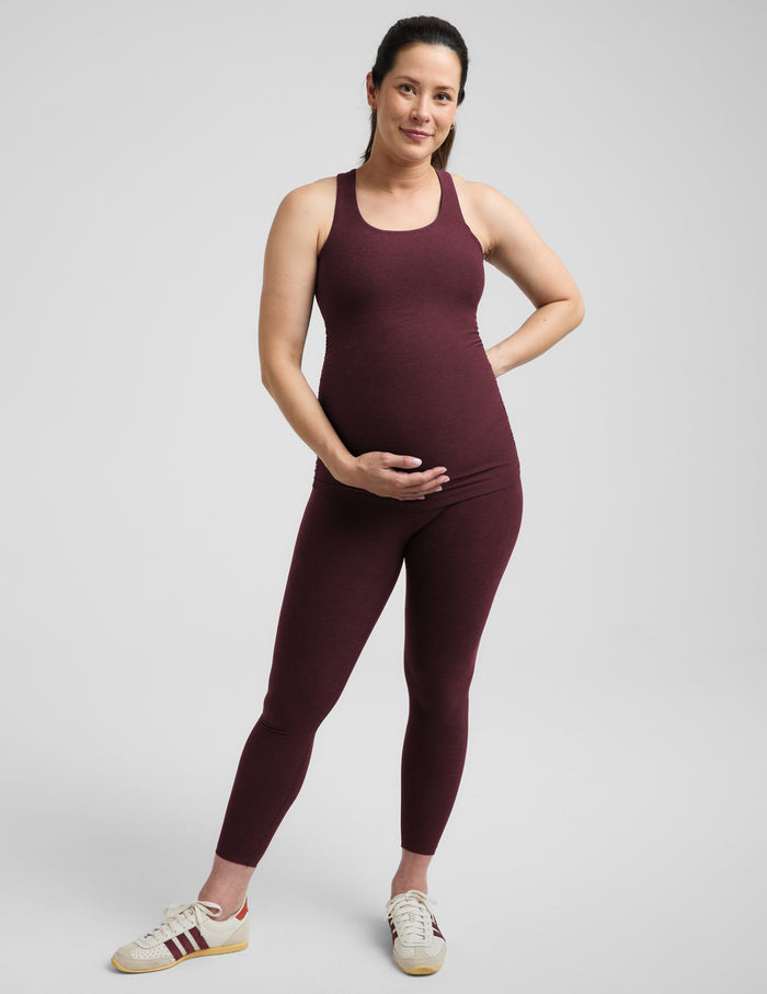 A pregnant woman wearing a matching dark burgundy maternity tank top and leggings set, standing against a solid light gray background.