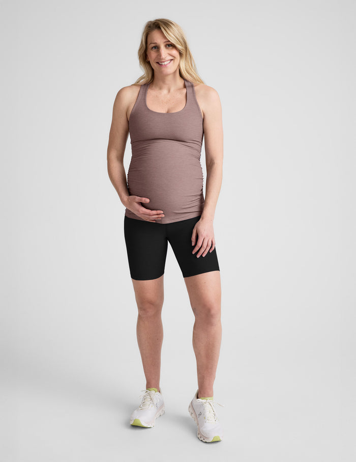Pregnant woman wearing a brown tank top and black shorts on a light gray background.