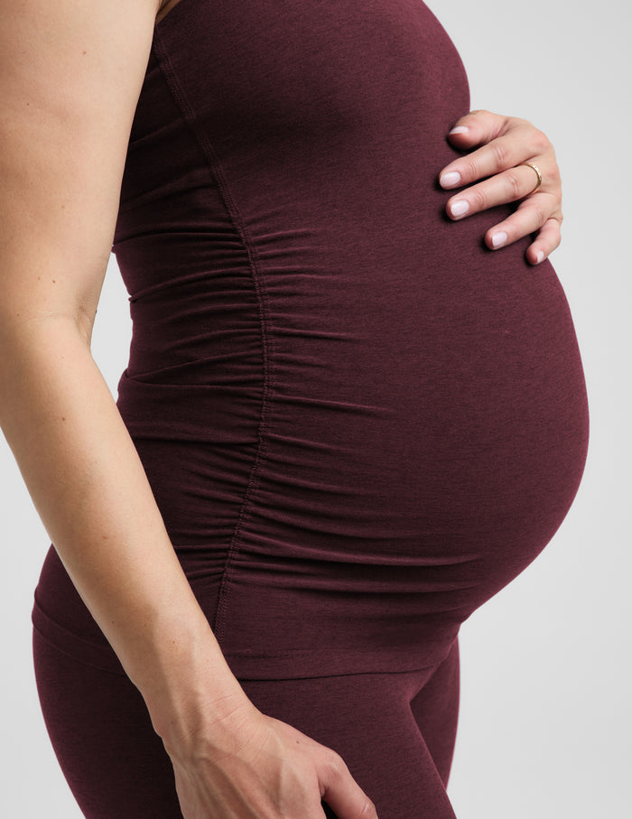 A pregnant woman wearing a matching dark burgundy maternity tank top and leggings set, standing against a solid light gray background.
