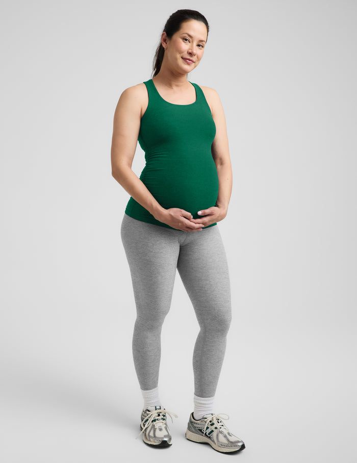 Pregnant woman wearing a green maternity racerback tank and gray leggings against a plain background.