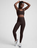 woman wearing brown high-waisted midi length leggings with pockets on a plain background.