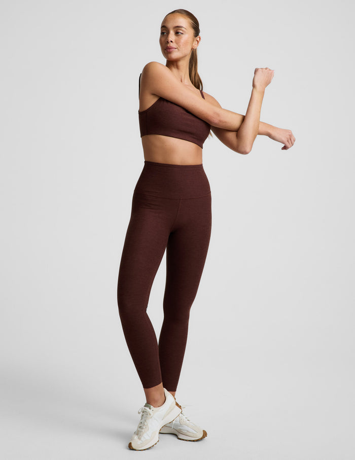Woman wearing a brown sports bra and brown high-waisted midi length leggings against a plain background.