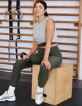 Woman in a grey cropped tank and green high-waisted midi length leggings sitting on a wooden box in a gym setting.