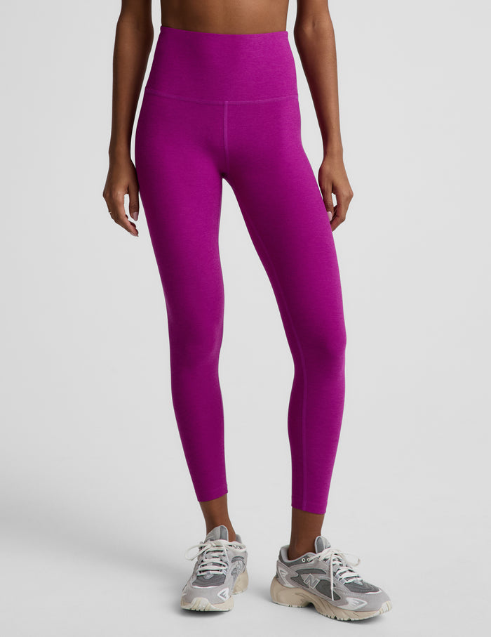 Woman wearing a purple bra and purple high-waisted midi length leggings against a plain background.