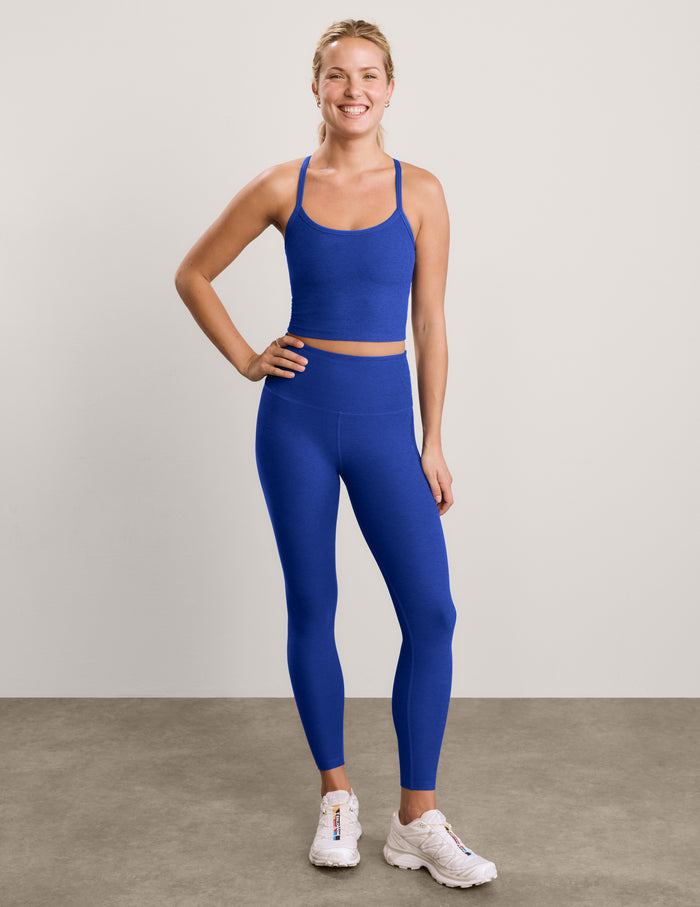 A woman modeling a matching royal blue activewear set consisting of a cropped tank and high-waisted leggings, paired with white sneakers, against a neutral studio backdrop.