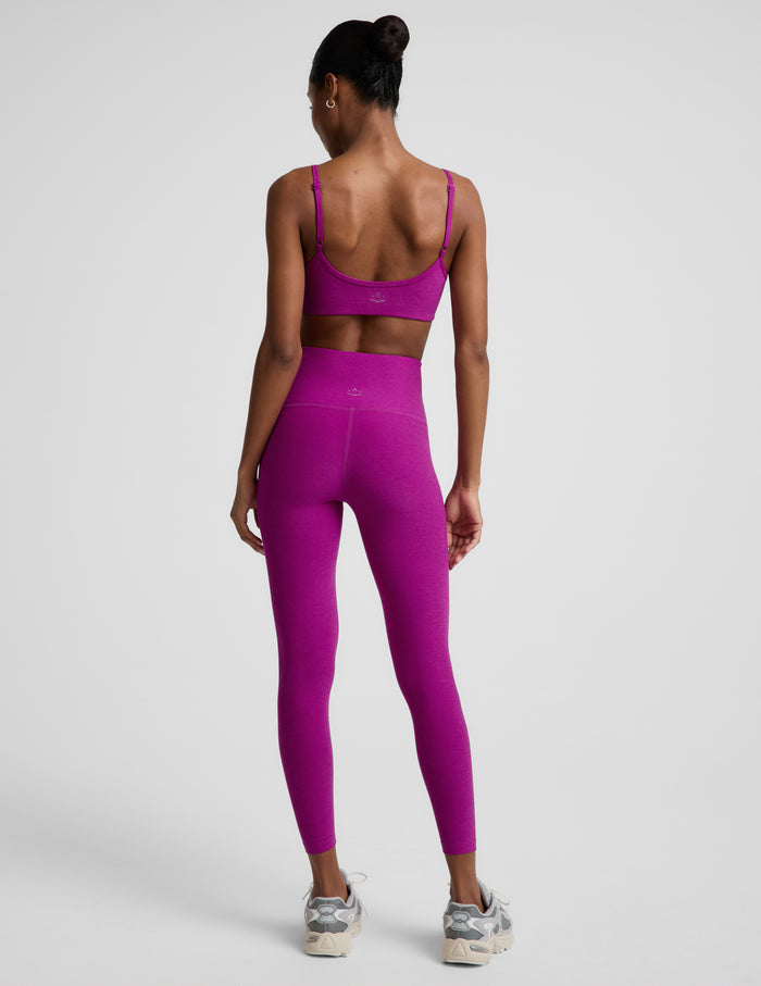 Woman wearing a purple bra and purple high-waisted midi length leggings against a plain background.