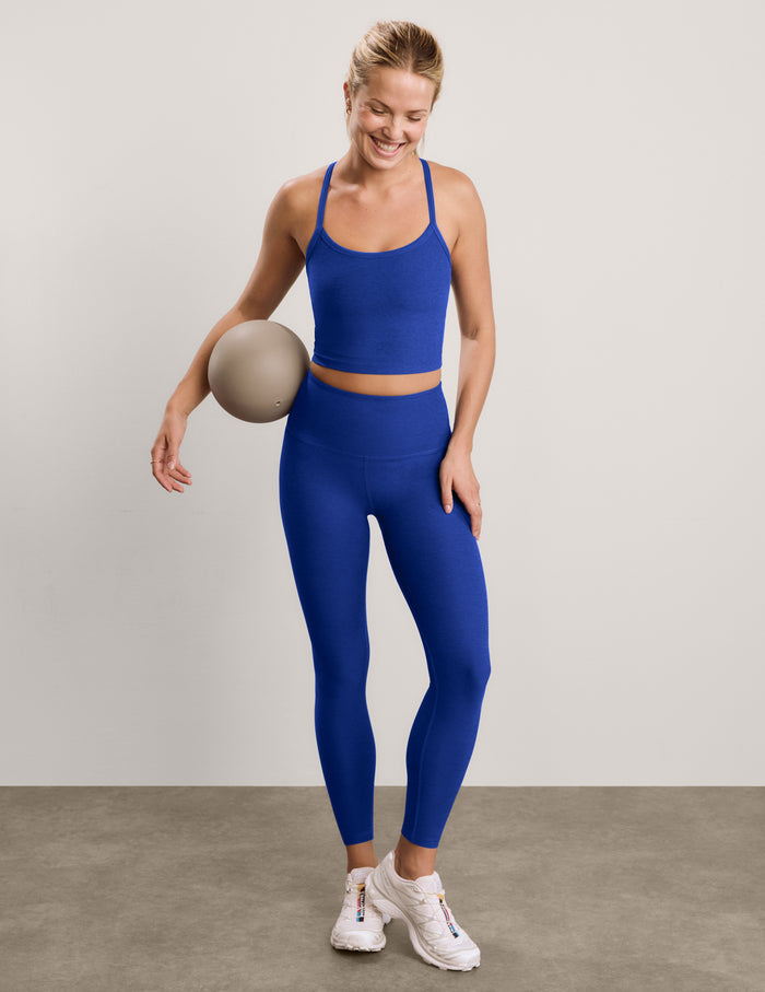 A woman modeling a matching royal blue activewear set consisting of a cropped tank and high-waisted leggings, paired with white sneakers, against a neutral studio backdrop.