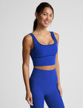 A woman modeling a matching royal blue activewear set consisting of a scoop-neck crop top and high-waisted leggings, photographed against a solid light gray studio background.